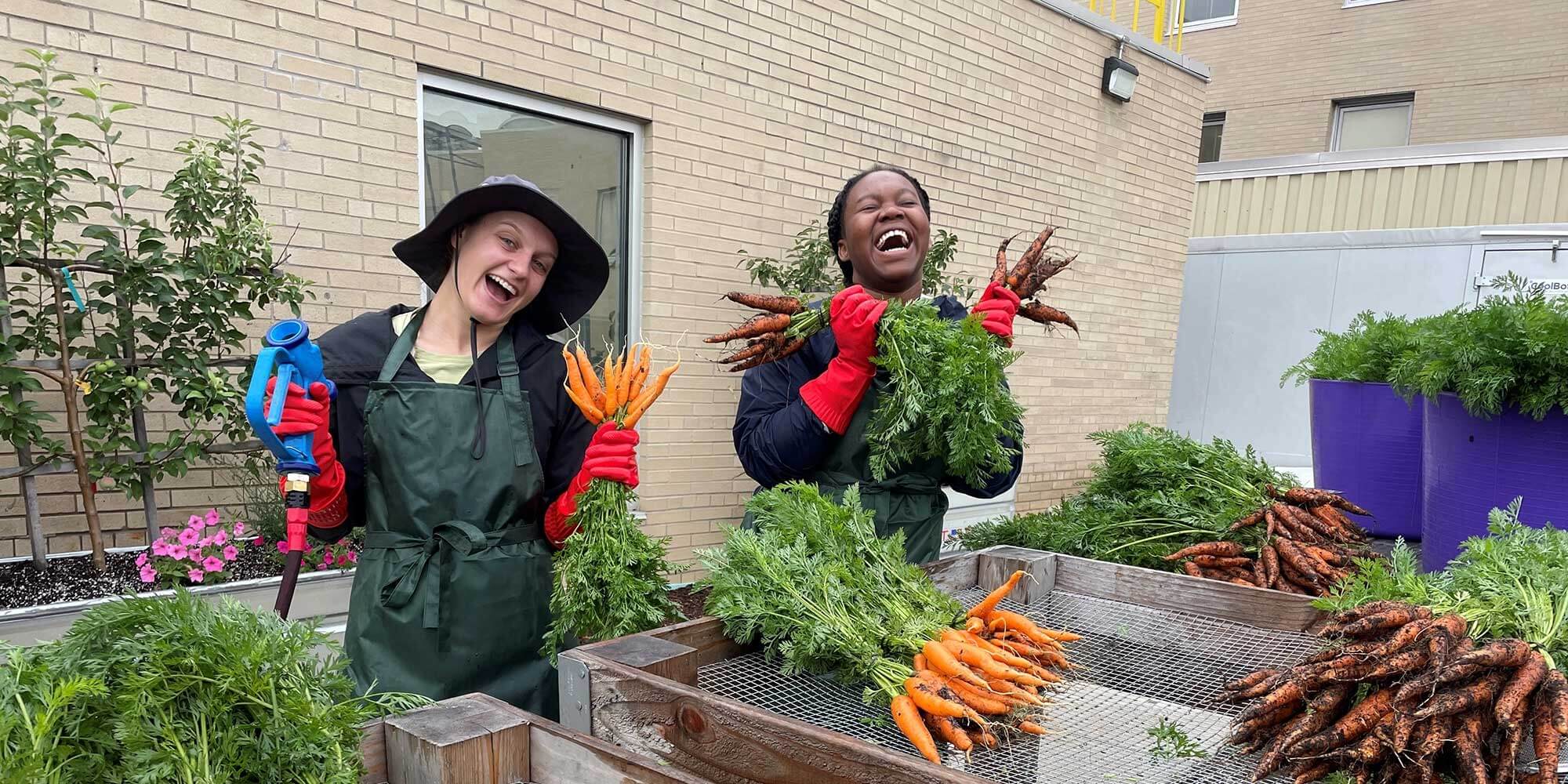Two women hold bundles of vegetables while laughing and joking together