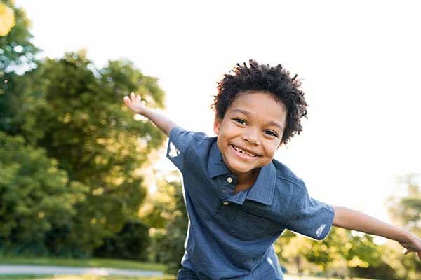 A smiling young boy runs outside in a park with his arms outstretched