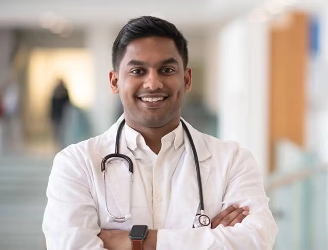 A male doctor stands with his arms crossed smiling in a hospital hallway