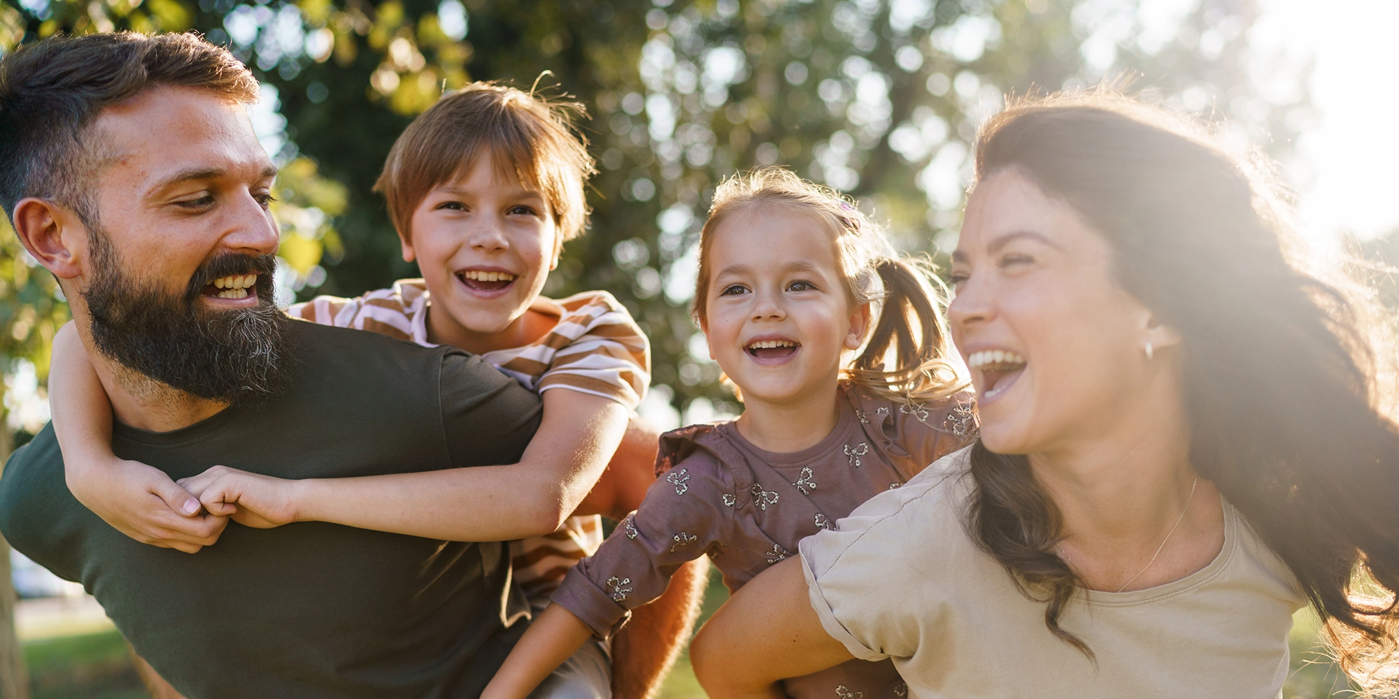 A mother and father carry their two children on their back. They're in a park; the sun is beaming in from behind them through the trees.