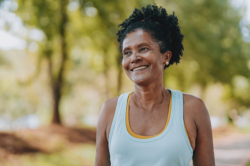A mature woman in a blue athletic tanktop smiles outside in a park