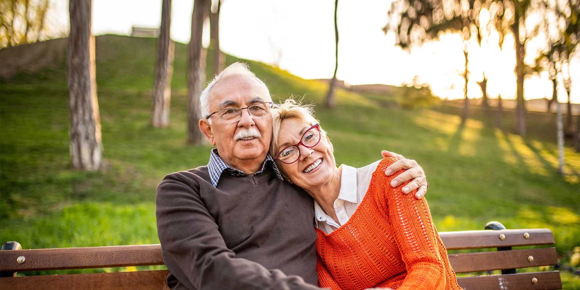 A senior couple embraces on a park bench. It's chilly outside and they're both wearing sweaters.
