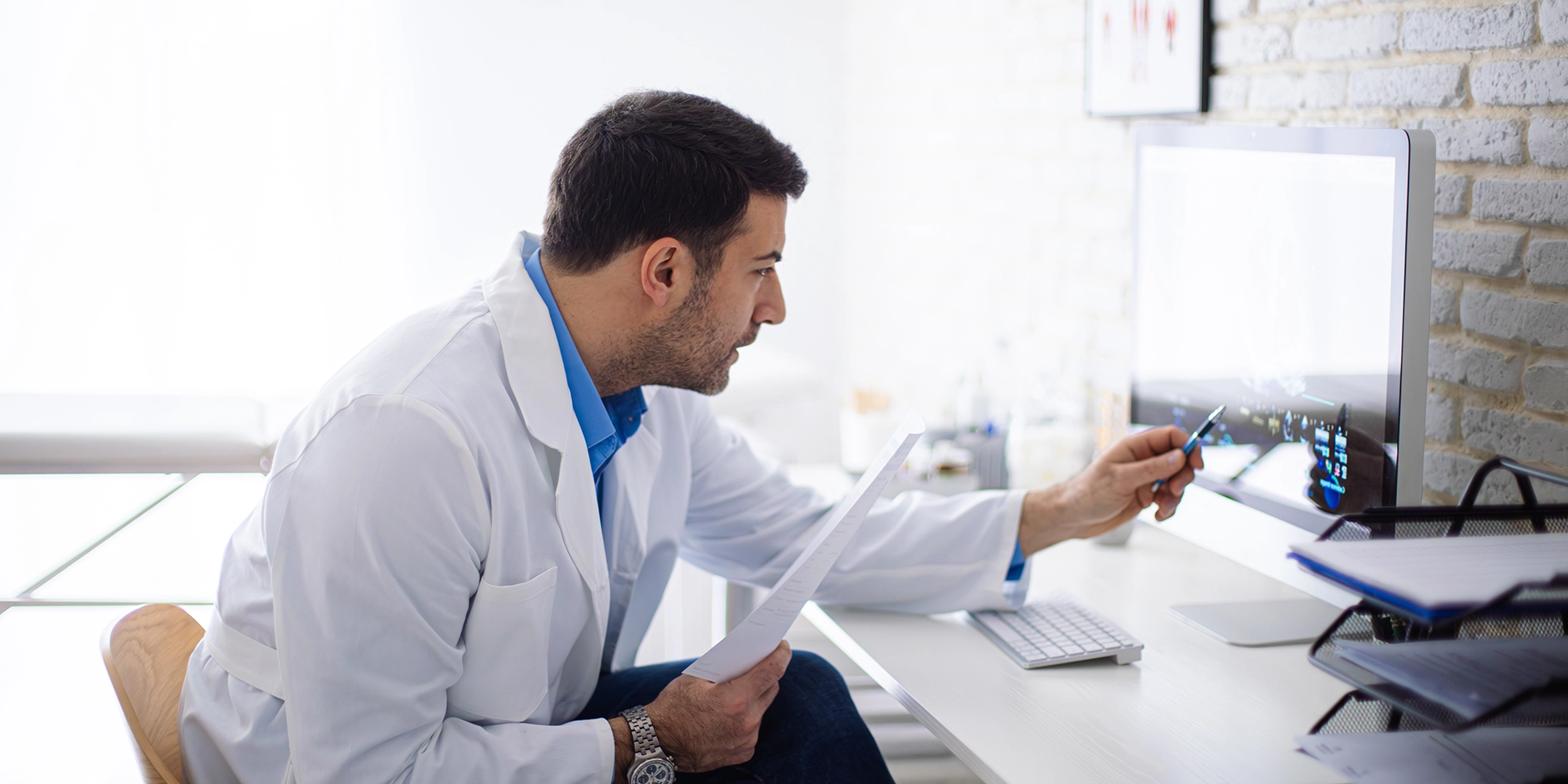 A male doctor leans in toward his monitor and taps his screen with a pen