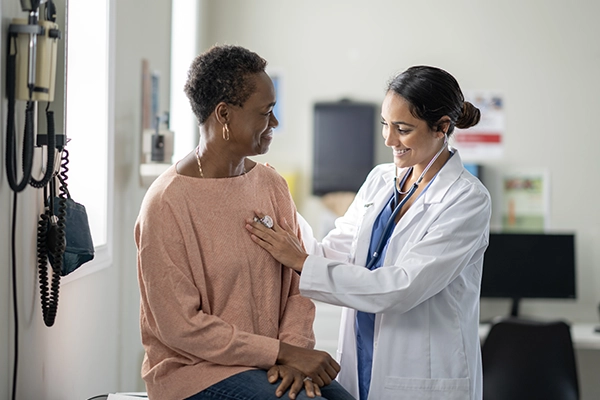 A woman meets with her primary care provider. The doctor is using a stethoscope on her.