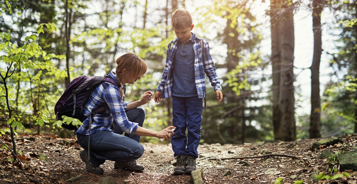 Mother sprays young son with insect repellant while on a hike in wooded forest.