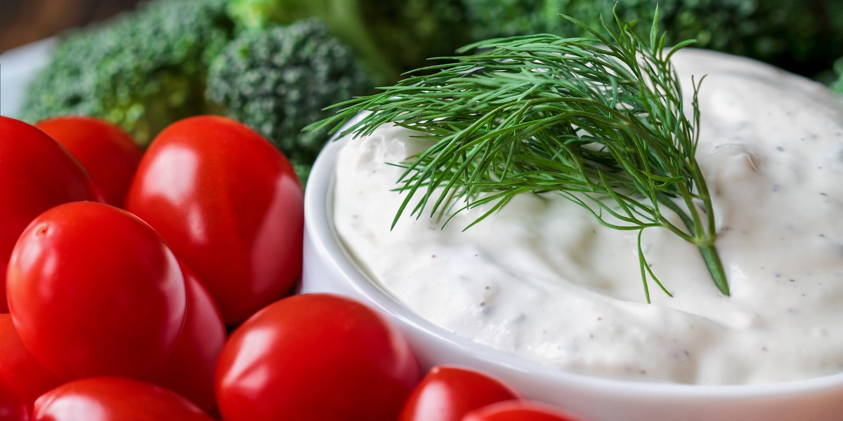 Creamy ranch dressing in a white bowl with a dill weed garnish surrounded by fresh tomatoes and broccoli.
