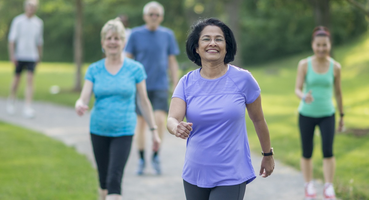 Mature woman wearing a purple exercise shirt walking outside with other people in a park.