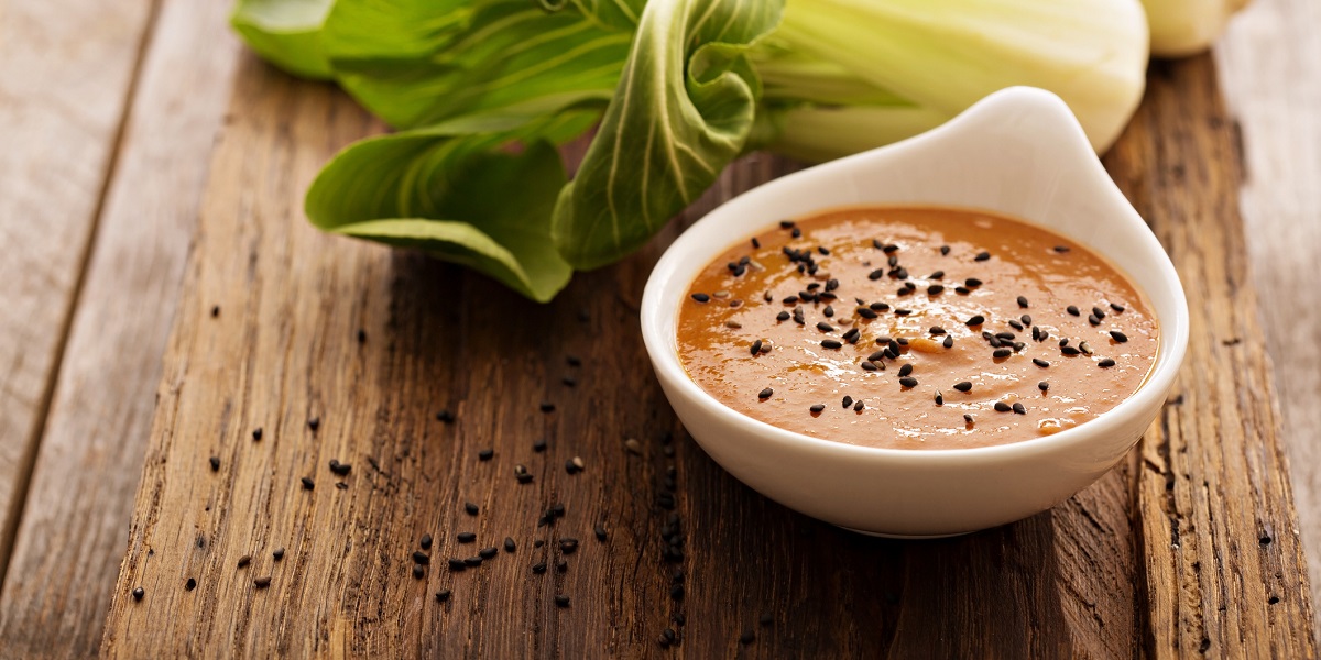 Ginger dressing sprinkled with black sesame seeds in small white bowl on aged wooden table with bok choi in background.