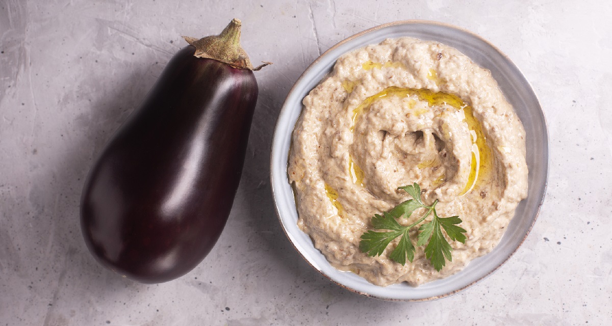 One ripe purple eggplant next to a bowl of creamy eggplant dip with a parsley garnish, both on top of a marble countertop.